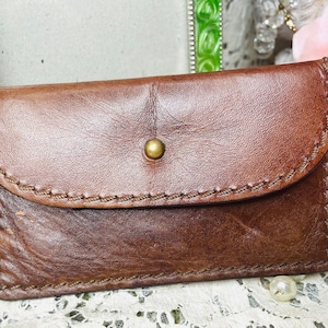 May include: A rectangular brown leather card holder featuring a curved flap closure secured with a single brass-toned metal stud. The edges of the leather are finished with visible, thick brown stitching that outlines the entire perimeter.