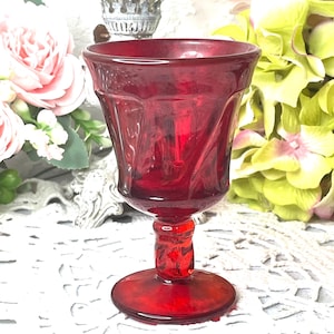 May include: A ruby red vintage goblet with a detailed stem and a round base. The glass has a textured design and a flared rim. The goblet is displayed in front of a backdrop of flowers.