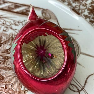 May include: A vintage Christmas ornament with a deep red, teardrop-shaped body. The ornament features a central, reflective gold-colored design and is accented with silver and green leaf-like details. The ornament is likely made of glass.