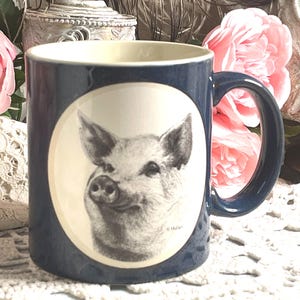 May include: A dark blue ceramic mug featuring a central cream-colored oval medallion. Inside the oval is a detailed black and white sketch of a pig's head, with the small signature V. Muller visible near the bottom right of the pig illustration.