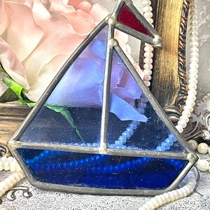 May include: A stained glass sailboat pendant with a red flag, blue sail, and dark blue base. The sailboat is framed in silver and features a beaded accent. The background includes a pink rose and pearl necklaces.