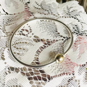 May include: A circular silver bangle bracelet featuring a single polished gold-tone spherical bead accent. The metal band is smooth and continuous, showcasing a metallic contrast between the silver-colored hoop and the gold-colored bead.
