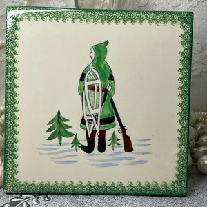 May include: A square ceramic tile with a cream-colored background and a green decorative border. The illustration depicts a figure in a green hooded coat carrying snowshoes and a rifle, standing in a snowy landscape with small pine trees.