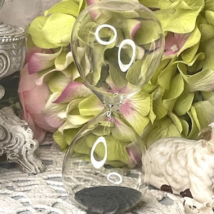 May include: Clear glass hourglass with white circular accents. The hourglass contains dark gray sand. The hourglass is set against a backdrop of pink and green flowers and a white lace surface.