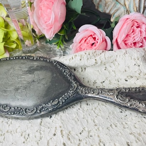 May include: Antique silver-toned hand mirror with an oval mirror and ornate handle. The mirror's frame and handle feature detailed scrollwork. The background includes pink roses and lace.