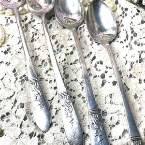 May include: A close-up of four silver spoons with ornate floral designs on the handles. The spoons are arranged diagonally, showcasing their polished surfaces. The spoons rest on a white lace tablecloth.