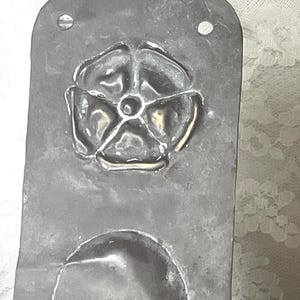 May include: A vintage, rectangular metal mold with a raised floral design and a curved indentation. The metal has a dark, aged appearance, with visible wear and tear. Two small holes are present near the top edge.