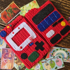 Op de afbeelding: Een rood gehaakt etui ontworpen als een draagbare spelcomputer, voorzien van een wit vilt schermkader, een zwart vilt D-pad en diverse blauwe, zwarte, witte en gele viltknopaccenten op de voor- en binnenpanelen.