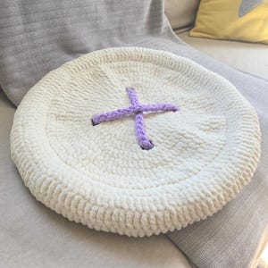 May include: A round, cream-colored crocheted cushion shaped like a large button. The cushion features a thick, raised border and a central X-shaped detail made of two intersecting strands of light purple yarn, mimicking the appearance of button holes.