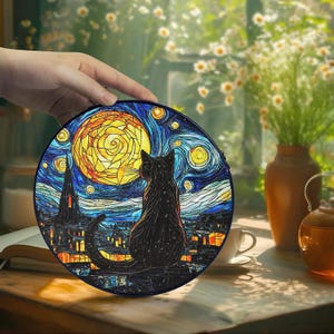 May include: A round, black cat-themed decorative item featuring a stained-glass style design. The image depicts a black cat gazing at a yellow moon against a starry night sky, with a town silhouette. The item is held by a hand.