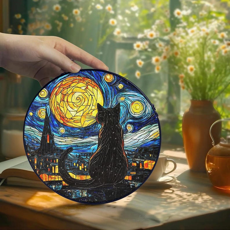 May include: A round, black cat-themed decorative item featuring a stained-glass style design. The image depicts a black cat gazing at a yellow moon against a starry night sky, with a town silhouette. The item is held by a hand.