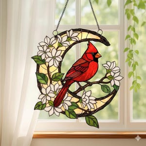 May include: Stained glass window hanging featuring a vibrant red cardinal perched on a branch with white flowers and green leaves. The bird is set against a yellow background within a crescent-shaped frame, suspended by a chain.