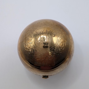 May include: A gold-coloured, round compact with floral and line engravings. The top features an ornate monogram within a floral design. The compact has a small clasp and a ridged edge, suggesting a vintage or antique style.