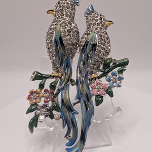 Puede incluir: Broche decorativo con dos pájaros enjoyados posados en una rama con flores coloridas. Los pájaros tienen cuerpos plateados cubiertos de pequeños cristales, con plumas de cola azules y verdes. La rama y las flores están pintadas con esmalte.