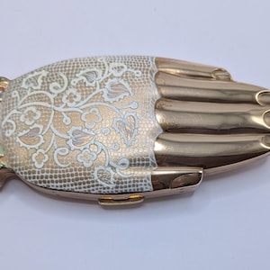 Peut inclure: Une boîte à bijoux en forme de main dorée. La paume présente un motif de dentelle florale crème. Le poignet a une bordure décorative avec des accents colorés. Les doigts sont lisses et détaillés.