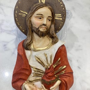 Può includere: Un'immagine religiosa da parete raffigurante un busto di Gesù Cristo. La scultura presenta un'aureola marrone, lunghi capelli castani e una barba. Indossa una veste rossa e tiene in mano un cuore fiammeggiante. La scultura è in materiale chiaro.