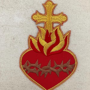 Könnte beinhalten: Ein gestickter Aufnäher mit einem roten Herz, das von einer braunen Dornenkrone umgeben ist. Über dem Herz befinden sich goldgelbe Flammen und ein goldgelbes Kreuz. Das gesamte Motiv ist mit einem dünnen goldgelben Rand auf rotem Stoff versehen.