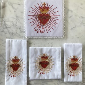 Può includere: Lenzuola bianche ricamate con un disegno del Sacro Cuore. Il cuore è rosso con una corona e raggi dorati, alcuni hanno bordi in pizzo. Le lenzuola sono di varie dimensioni, tra cui una forma quadrata e rettangolare.