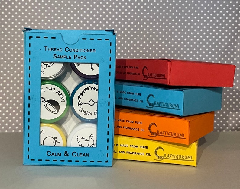 May include: A blue box with a clear window showing six small round containers of thread conditioner. The containers have different colored lids and labels with various designs and text, including "Coconut", "Kingdom of", and "Calm & Clean". The box is labeled "Thread Conditioner Sample Pack".