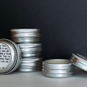 May include: Several small, silver-colored tins are stacked and arranged on a light surface against a dark background. One tin is open, and another has its lid slightly ajar. Text on a label describes how to use the product.