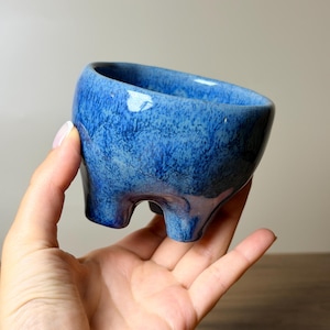 Könnte beinhalten: Ein kleiner, runder Keramikübertopf mit einer glänzenden, gesprenkelten blauen Glasur. Das Gefäß hat eine weite, offene Oberseite und wird von drei kurzen, stabilen Füßen an der Basis getragen, was eine feste Standfläche für kleine Pflanzen bietet.