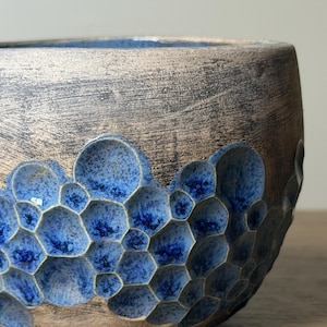 Puede incluir: Un cuenco de cerámica con un acabado exterior texturizado en tonos bronce metalizado. La parte inferior presenta un grupo de hendiduras en forma de panal, esmaltadas en tonos vibrantes de azul cobalto profundo y azul brillante.
