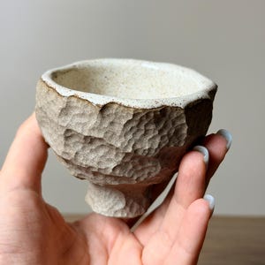 May include: A small, hand-held ceramic bowl with a textured, faceted exterior in a light beige tone. The interior features a smooth, speckled off-white glaze, while the rim has a dark brown, irregular edge that contrasts with the rough, clay-like surface.