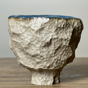 May include: A textured, beige ceramic bowl with a blue rim. The bowl has a rough, carved exterior and a small, stylized symbol etched into the side. The bowl sits on a small, pedestal-like base.