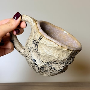 Pode incluir: Uma caneca de cerâmica artesanal com uma superfície exterior texturizada e irregular, apresentando um esmalte de cor creme e padrões de fissuras pretas proeminentes. A caneca tem uma pega grossa e curva e um rebordo interior liso e claro.