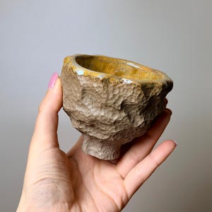 May include: A small, hand-held ceramic cup with a textured, dimpled exterior in a matte beige finish. The interior of the cup features a smooth, glossy, golden-yellow glaze that spills over the uneven, organic rim of the vessel.