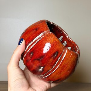 Può includere: Una ciotola in ceramica rotonda, realizzata a mano, con una smaltatura lucida di colore rosso e arancione vivace. La superficie presenta creste verticali strutturate e accenti scuri e maculati, con un'ampia apertura e finitura liscia.