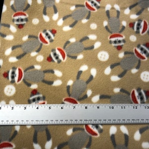 May include: Tan fabric with a repeating pattern of gray sock monkeys wearing red hats. The fabric is measured with a ruler in inches.