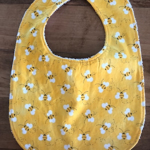 Puede incluir: Un babero amarillo para bebé con un patrón de abejas blancas. El babero tiene un cierre de presión blanco.