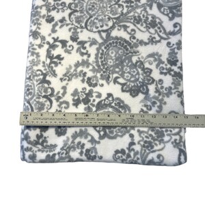 Puede incluir: Una manta de forro polar blanca y gris con un patrón de paisley. La manta mide aproximadamente 38 cm de ancho y 41 cm de largo.
