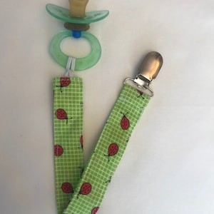 May include: A green and white pacifier clip with a ladybug print. The clip has a silver metal clasp and is attached to a green pacifier with a clear silicone ring.