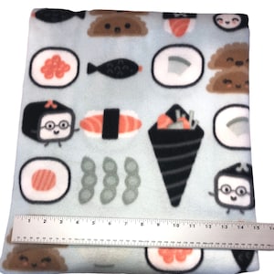 Op de afbeelding: Een lichtblauwe fleece deken met een herhalend patroon van sushi, edamame en vis. De sushi is afgebeeld als cartoonfiguren met gezichten. De deken is 38 cm lang.