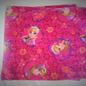 May include: Pink fleece blanket featuring a repeating pattern of a cartoon girl with blonde hair and blue eyes. The girl is wearing a blue and purple dress and a crown. The blanket has a white background with a pink and purple floral pattern.