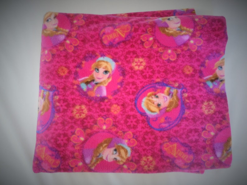 May include: Pink fleece blanket featuring a repeating pattern of a cartoon girl with blonde hair and blue eyes. The girl is wearing a blue and purple dress and a crown. The blanket has a white background with a pink and purple floral pattern.