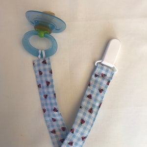 May include: A blue and white gingham pacifier clip with a ladybug print. The clip has a white plastic clip and a blue pacifier holder.