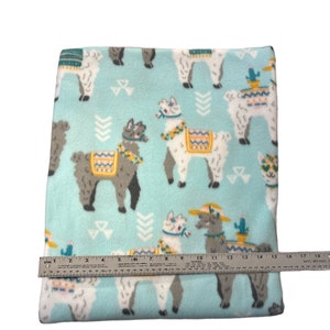 May include: A light blue fleece blanket with a pattern of llamas wearing hats and carrying cacti. The blanket is approximately 17 inches wide.
