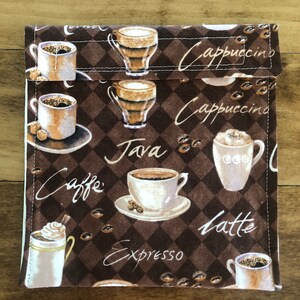 Op de afbeelding: Een bruine stoffen lunchtas met een geruit patroon en verschillende koffie-thema illustraties. De tas heeft de woorden "Cappuccino", "Caffe", "Java", "Latte" en "Espresso" erop gedrukt.