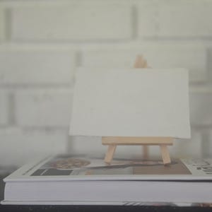 May include: A small, light-coloured wooden easel holding a blank, rectangular white canvas. The easel is positioned on top of a closed book with a white cover, featuring a visible spine and a printed image on the front cover surface.