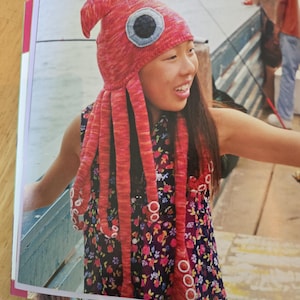 Puede incluir: Un gorro de punto con forma de calamar en tonos rojos y naranjas, con un gran parche circular para el ojo y largos tentáculos colgantes con detalles de ventosas circulares blancas. El gorro está hecho de hilo texturizado y es puntiagudo.