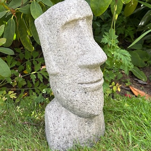 May include: A grey stone-textured planter shaped like a Moai head statue. The sculpture features a prominent nose, a defined brow ridge, and a subtle horizontal line for the mouth. The top of the head is open, creating a hollow space for plants.