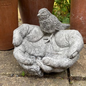 May include: A grey stone sculpture featuring two cupped hands holding a small bird. The textured surface of the sculpture shows detailed finger and palm contours, with the bird perched centrally on the palms. The material has a weathered finish.