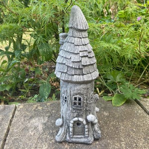 May include: A grey stone garden ornament shaped like a tall, slender tower with a textured shingled roof, a small arched door, and a window. The structure features a bark-like exterior finish with small mushroom-shaped accents at the base.