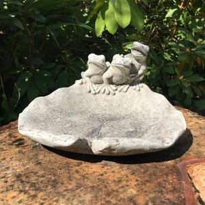 May include: A grey stone birdbath with three frog figurines on the rim. The birdbath has a shallow, leaf-shaped basin. The background features green foliage and is set on a wooden surface.