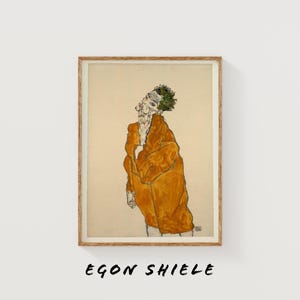 May include: Framed artwork featuring a drawing of a person in a mustard-colored coat. The artwork has a minimalist style with black outlines and a neutral background. The text "EGON SHIELE" is printed in black at the bottom of the frame.