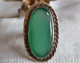 May include: An oval ring featuring a smooth, light green cabochon stone set within a twisted metal border. The setting has a metallic finish that frames the central green stone, creating a distinct oval shape for the piece of jewelry.