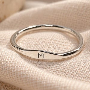 Op de afbeelding: Een dunne, gepolijste zilverkleurige ring met een plat, horizontaal gedeelte aan de voorzijde. In het midden van de platte balk is de hoofdletter M gegraveerd in een eenvoudig, strak lettertype. De ring heeft een gladde, ronde vorm.